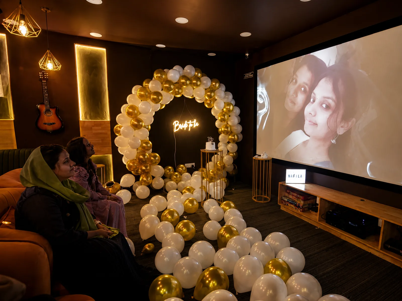 Inside ShowTime Max: guests on the tan sofa facing the projection screen, with a white and gold balloon ring beside it