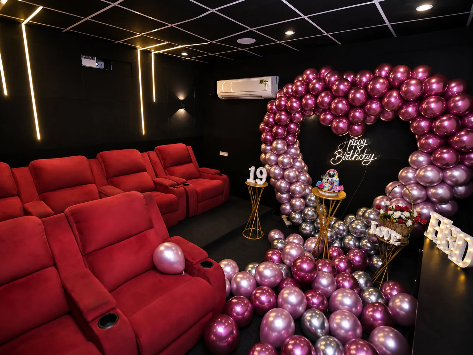 The same room set up for a birthday — a pink and chrome heart arch, a neon Happy Birthday sign, a lit 19 and a cake on a gold stand
